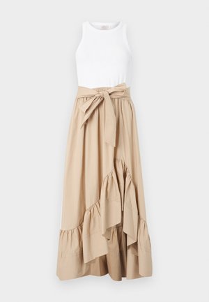 Naine kannab valget varrukateta topi, pruunikat asümmeetrist volditud seelikut vööga ja beežid tossusid, seisab ühevärvilise halli seina taustal.