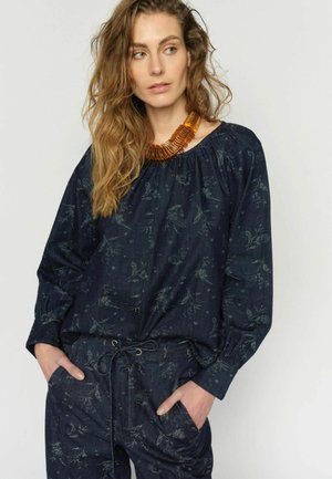 Donna con capelli mossi indossa una maglia a maniche lunghe blu marina con fiori e pantaloni coordinati, accessoriata con una collana spessa color ambra, mani in tasca.