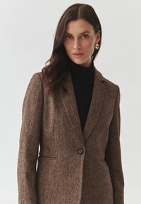 Blazer maro, cu textură, cu închidere printr-un singur nasture, rever ascuțit și o croială ajustată, purtat peste un pulover negru cu guler înalt. Cercei aurii vizibili.