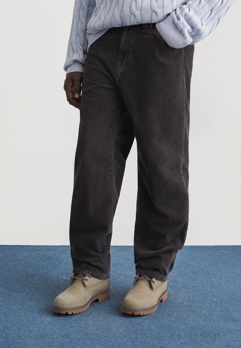 Personne portant un pantalon en velours côtelé noir, un pull en tricot torsadé gris clair et des bottes beiges à lacets, debout sur un tapis bleu.