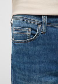 Detail vrecka modrých denimových džín s detailným prešitím a béžovou košeľou zastrčenou v páse.