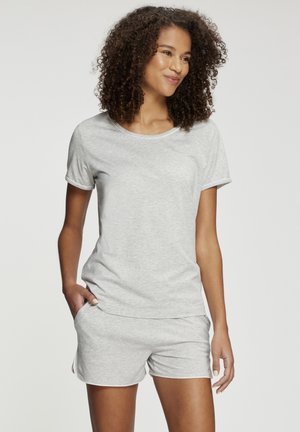 Woman with curly hair wearing light gray short-sleeve shirt and matching shorts, standing with one hand in pocket against a plain background.