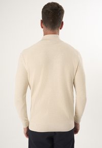 Beżowy sweter z dzianiny o prążkowanej fakturze, z długimi rękawami i wysokim kołnierzem. Projekt charakteryzuje się dopasowanym krojem i subtelną elegancją.