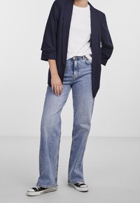 Marinblazer över en vit t-shirt, i kombination med ljusblå utsvängda jeans och svarta kanvas sneakers. Märkbar sömnadsdetaljering på jeansen.