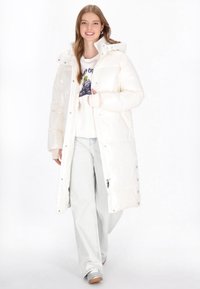 Hosszú, fehér puffernadrág, kapucnival, elöl patentgombokkal, fényes textúrával, grafikus kapucnis pulóver és világos farmernadrág fölött viselve.