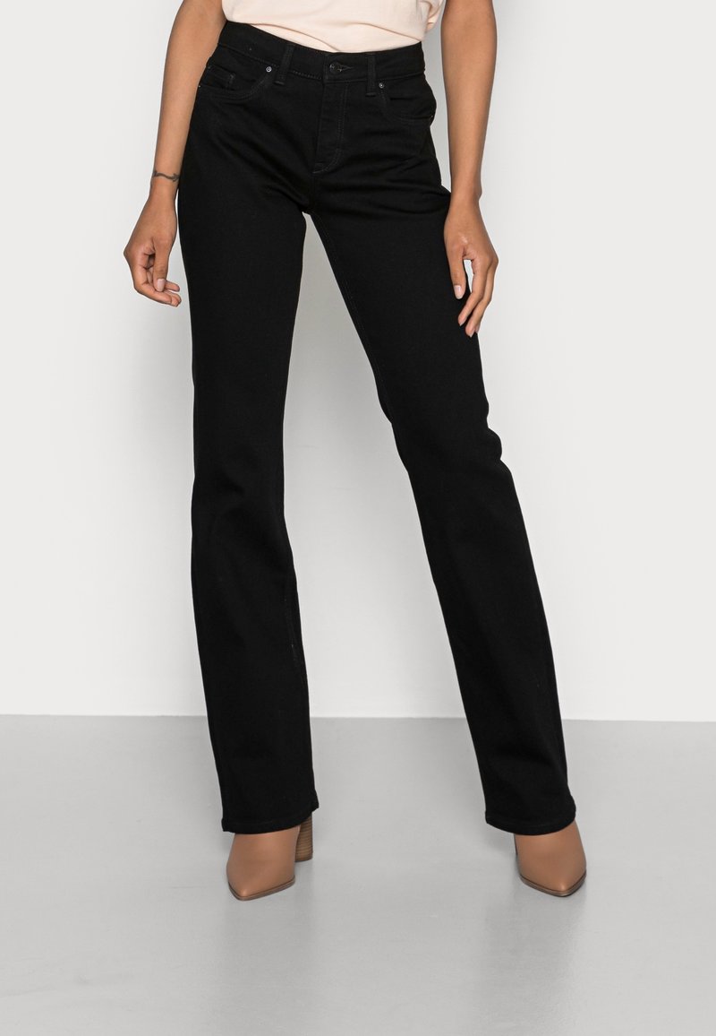 Femme portant un jean noir taille haute, coupe droite, et des bottines beiges à talons pointues, debout devant un fond clair uni.
