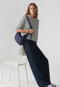 Gestreepte, korte mouwen top in wit en marineblauw, gecombineerd met wijde marineblauwe broek. Een marineblauwe schoudertas hangt over de arm. Minimalistische setting.