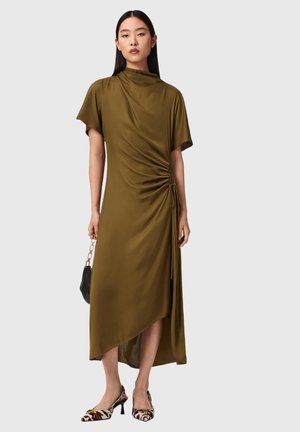 Kvinde iført olivengrøn asymmetrisk midi-kjole med samlet detalje i siden, der holder en sort håndtaske og har på dyreprintede hæle foran en ensfarvet baggrund.