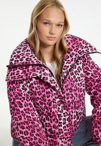 Růžová pufová bunda s leopardím potiskem, vysokým límcem, zapínáním na zip a lesklou texturou. Má velké černé skvrny a volný střih.