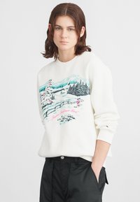 Sweatshirt branco com uma gráfica de paisagem de inverno, apresentando uma árvore rosa e montanhas, em contraste com calças pretas de corte relaxado.