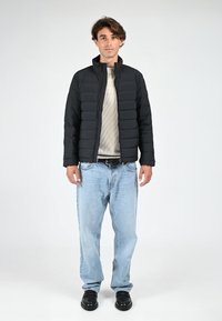 Veste rembourrée noire matelassée avec un col montant, portée par-dessus un pull en maille beige, associée à un jean bleu clair et des chaussures noires.