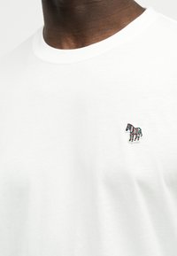 Primer plano de una persona que lleva una camiseta blanca con una pequeña cebra a rayas de colores bordada en el pecho izquierdo.