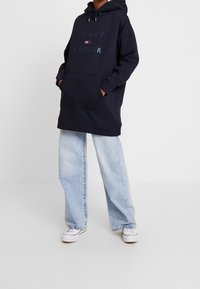 Sudadera azul marino con logotipo en lettering metálico, que cuenta con un bolsillo tipo canguro, capucha y un corte holgado, combinada con unos pantalones vaqueros de pierna ancha en azul claro.