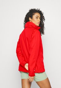 Rød zip-up jakke med høy krage, justerbare ermer og en lett tekstur. Båret over lysegrønne shorts.