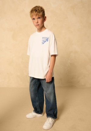 Vit kortärmad t-shirt med blå grafisk text; tillsammans med lösa blå jeans och vita sneakers. Neutral bakgrund framhäver fokus.