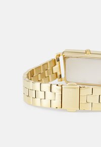 Orologio da polso in oro con cassa rettangolare e bracciale in metallo a maglie. Presenta un design minimalista e una superficie liscia e riflettente.