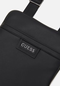 Schwarze synthetische Leder-Umhängetasche mit strukturiertem Finish, ausgestattet mit einem auffälligen silbernen "GUESS"-Logo und verstellbarem schwarzen Trageriemen.