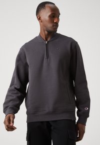 Sudadera gris de tejido suave con medio cierre en el cuello, puños acanalados y un pequeño logo en el pecho. Tiene un corte holgado y una textura suave.