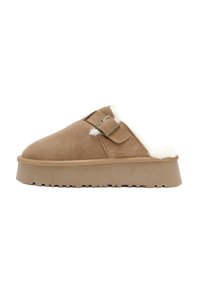 Bruine suede instappers met een witte faux bont voering, een platte zool en twee gesps aan de bovenkant. Zachte textuur overal.