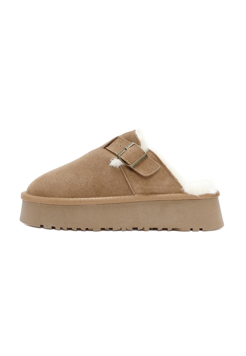 Bruine suede instappers met een witte faux bont voering, een platte zool en twee gesps aan de bovenkant. Zachte textuur overal.