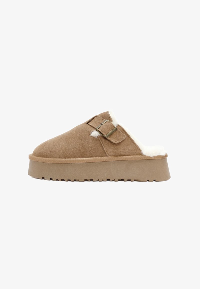 Bruine suede instappers met een witte faux bont voering, een platte zool en twee gesps aan de bovenkant. Zachte textuur overal.