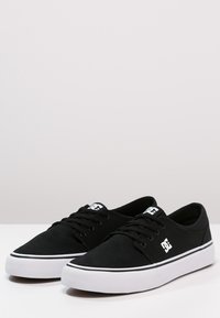 Black canvas sneakers with white rubber soles, featuring black laces, a contrasting white stripe, and logo on the side.