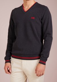 Dunkelgrauer V-Ausschnittpullover aus weichem Material, mit rotem Saum am Kragen und den Bündchen, sowie einem roten Logo auf der Brust.