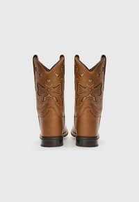 Stivali da cowboy in pelle marrone con intagli decorativi a forma di cuore e fiocco. Presentano un colletto appuntito e suole in gomma nera.