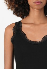 Svart ribbet tanktopp med en bølget blonderkant rundt halslinjen og skuldrene, med en myk tekstur og kroppsformet design.