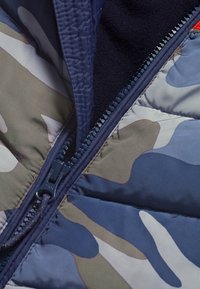 Kamelové zateplené bunda s maskáčovým vzorem, vybavená zipovým uzávěrem, tmavomodrou podšívkou a kombinací zelených, modrých a šedých tónů s hladkým, texturovaným povrchem.