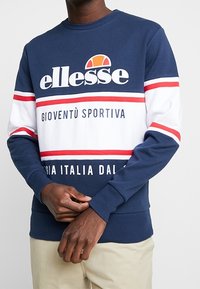 Man som bär en navy blå och vit ellesse-tröja med röda ränder och texten "GIOVENTÙ SPORTIVA" samt beige byxor, händerna knäppta vid midjan.