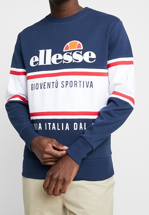 Man som bär en navy blå och vit ellesse-tröja med röda ränder och texten "GIOVENTÙ SPORTIVA" samt beige byxor, händerna knäppta vid midjan.