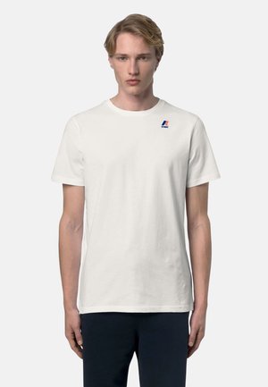 Giovane uomo con capelli biondi corti, indossa una semplice t-shirt bianca K-Way e pantaloni scuri, in piedi davanti a uno sfondo neutro.