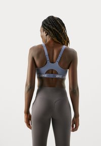 Mujer con el cabello trenzado, vistiendo un sujetador deportivo azul de Nike y leggins gris de cintura alta, de pie y mirando hacia atrás sobre un fondo blanco.
