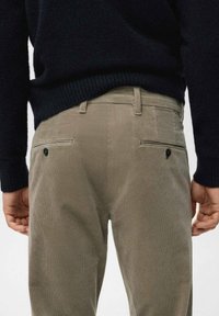 Man draagt beige corduroy broek met achterzakken met knopen en een zwarte trui, gefotografeerd van taille tot onder, staand tegen een effen achtergrond.