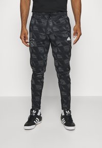 Pantalons de sport noirs avec un motif géométrique, dotés d'une taille élastique avec cordon de serrage, et accents du logo Adidas sur les côtés. Portés avec des baskets noires.