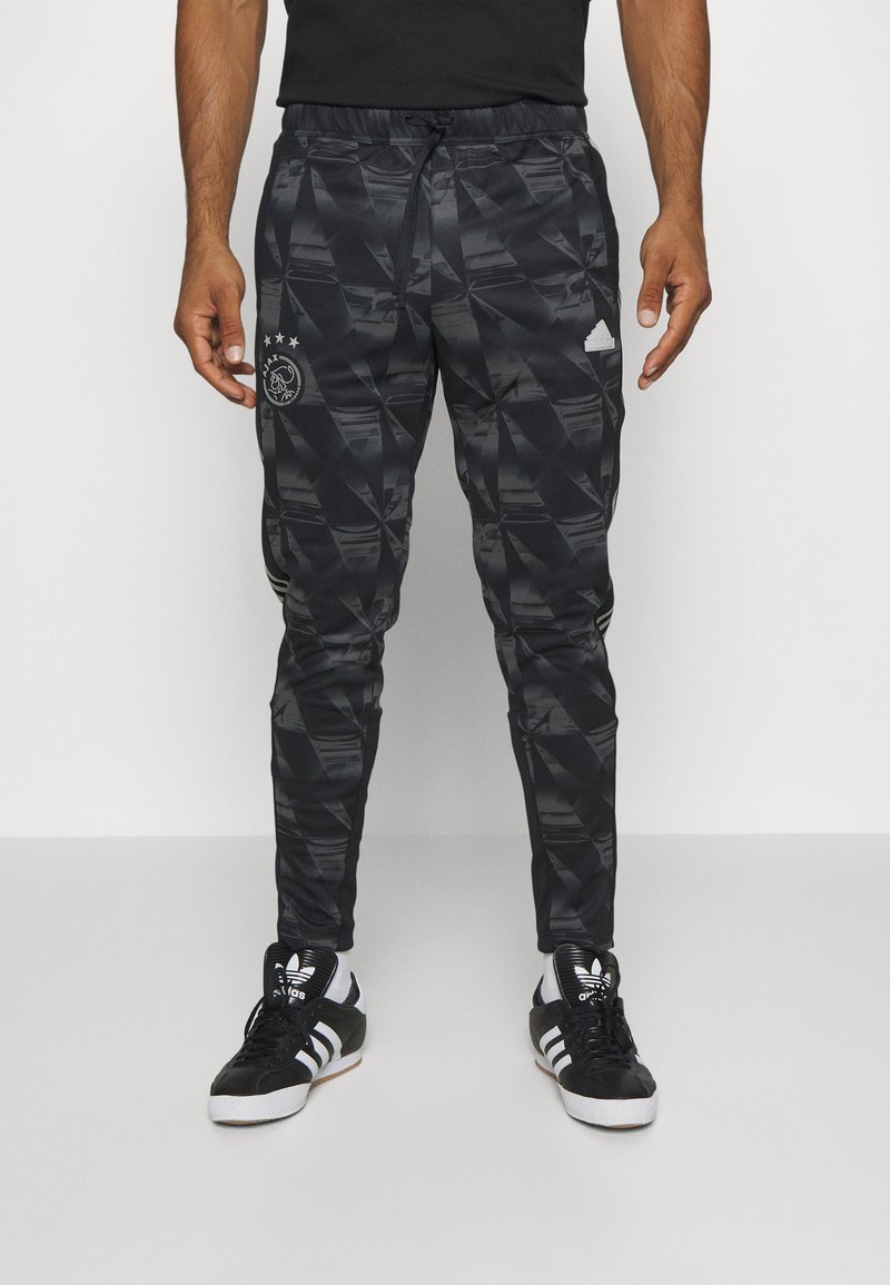Pantalons de sport noirs avec un motif géométrique, dotés d'une taille élastique avec cordon de serrage, et accents du logo Adidas sur les côtés. Portés avec des baskets noires.