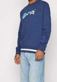 Man i marinblå sweatshirt med turkos och vit "AIR"-logotyp och ljusblå jeans mot enfärgad bakgrund.