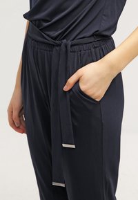 Pantalons larges bleu marine fabriqués dans un tissu lisse, dotés d'une taille élastique, de poches latérales et d'un détail de ceinture en tissu nouée.