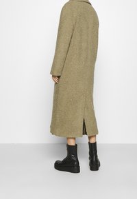 Lang, olivengrøn frakke med en tekstureret overflade og sidelukke, parret med sorte snørebåndsstøvler. Frakken har lange ærmer og en krave.