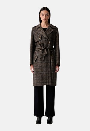 Donna che indossa un trench marrone a quadri con cintura sopra pantaloni neri e stivaletti neri, in piedi davanti a uno sfondo semplice.
