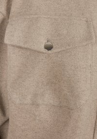 Poche en tissu beige avec un rabat sécurisé par un seul bouton rond argenté sur un vêtement en laine texturée.