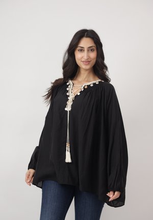 Kobieta o długich ciemnych włosach, ubrana w luźną czarną bluzkę z białymi frędzlami i niebieskie dżinsy, uśmiechająca się na jednolitym tle.