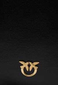 Χρυσό μεταλλικό στοιχείο δύο πουλιών αντίκρυ σε μέτωπο με ανοιγμένα φτερά, προσαρτημένο σε λείο μαύρο δερμάτινο επιφάνει�α.