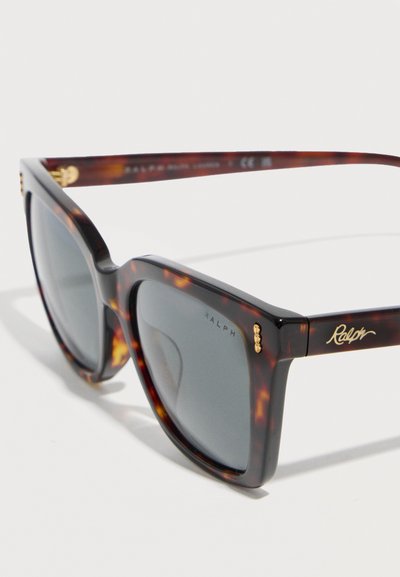 Γυαλιά ηλίου σε τετράγωνο σχήμα με χαβανέζικο μοτίβο, σκούρους φακούς, χρυσή αλυσίδα στο πλαίσιο και το λογότυπο "Ralph" στον βραχίονα.