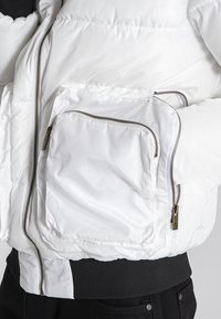 Giacca puffer bianca con finitura lucida, dotata di numerose tasche con zip e vita a coste nera, che mette in mostra una texture trapuntata.