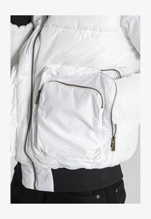 Giacca puffer bianca con finitura lucida, dotata di numerose tasche con zip e vita a coste nera, che mette in mostra una texture trapuntata.
