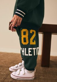 Green sweatshirt with white and navy striped cuffs, paired with green pants featuring yellow "82" and "ATHLETIC" print. White Velcro sneakers.