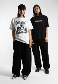 White graphic t-shirt with black print, paired with black baggy pants. The second person wears a black oversized t-shirt and black pants.