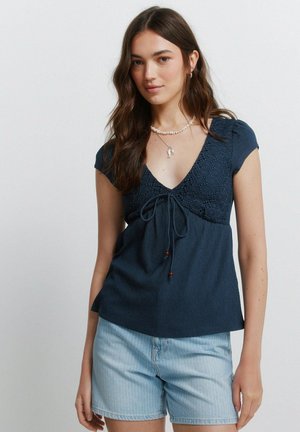 Ung kvinde med langt mørkt hår iført en marineblå tekstureret V-udskåret top og lyseblå denimshorts foran en ensfarvet baggrund.